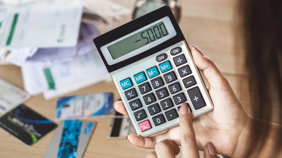 Kobieta trzymająca kalkulator z wynikiem -5000, na tle rachunków i kart płatniczych